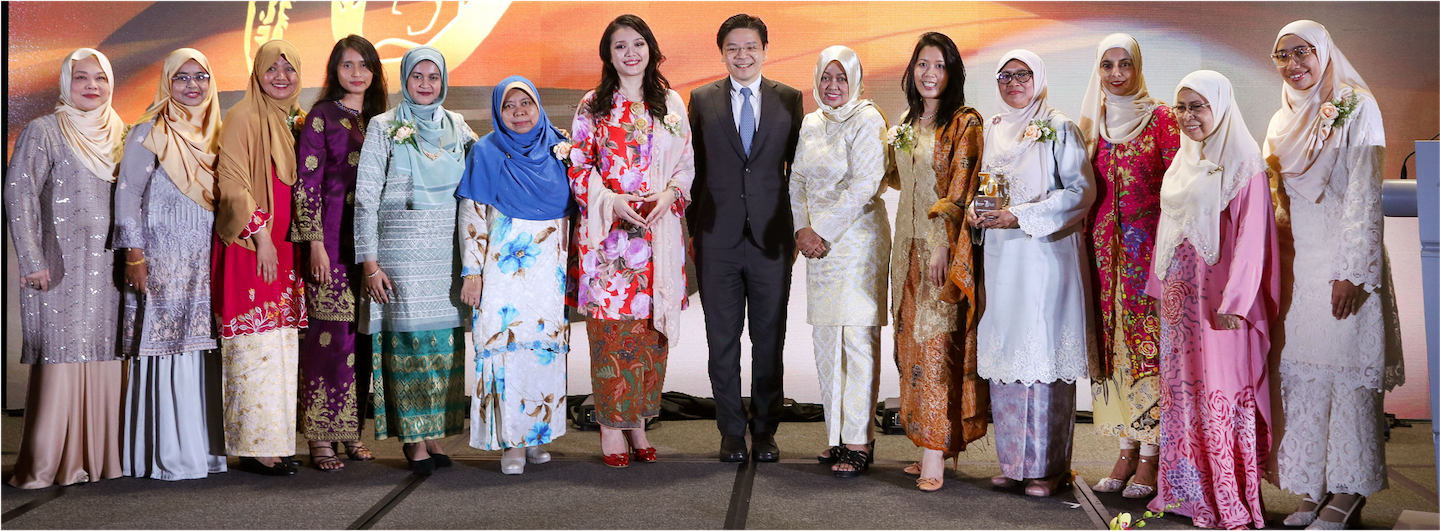 Group of women and a man in formal wear are posing on stage. Some women wear hijabs. One woman holds an award.